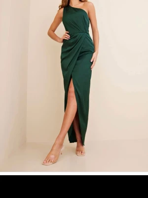 Mörkgrön oneshoulder långklänning - Elegant mörkgrön långklänning med oneshoulder-design och draperad omlott framtill. Klänningen har en hög slits som ger en snygg siluett och är perfekt för speciella tillfällen. Tillverkad i ett mjukt, följsamt material.