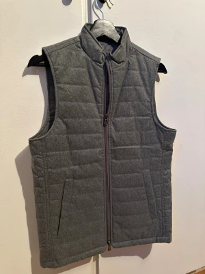 Mörkgrå quiltad dunväst - Stilren grå väst med quiltat mönster och hög krage. Västen har två snedställda fickor och stängs med dragkedja framtill. Perfekt för lager-på-lager. Strl XS.