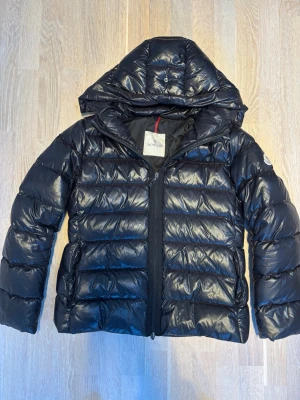 Svart dunjacka från Moncler med huva - Snygg svart dunjacka från Moncler med glansig finish och stor huva. Jackan har quiltad design, dragkedja framtill och Moncler-logga på ärmen. Perfekt för kalla dagar när du vill hålla stilen och värmen. Moncler storlek 3 vilket motsvarar M. 