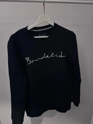 Svart sweatshirt från Bondelid - Snygg svart sweatshirt från Bondelid med vit broderad logga över bröstet. Klassisk rund halsringning och ribbade muddar vid ärmslut och nederkant. Perfekt för en avslappnad och stilren look.