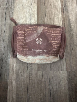 Brun necessär från The Body Shop - Necessär i brun och beige bomull från The Body Shop med textmönster och dragkedja. Tryck på framsidan med 'Exotically Creamy Coconut' och empowerment-ord i olika språk. Perfekt för smink eller småprylar.