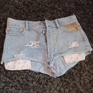 Jeansshorts - Snygga ljusblå jeansshorts från Ivyrevel med slitna detaljer. Framtill finns nitar i guld på ena fickan och fickorna som sticker ut har söta rosa små blommor🩵🌸✨️ Perfekta för sommaren! (OBS: De är ganska korta). Det är bara att skriva om man har frågor!😊💖