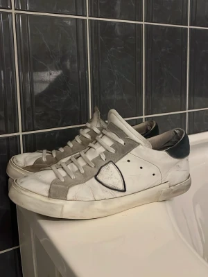 Vita Philippe Model sneakers med blå detaljer - Snygga vita sneakers från Philippe Model med blå mockadetaljer. Klassisk låg sommar sko. Är i bra skick förutom sittningen undertill men inget man märker av vid användning. 