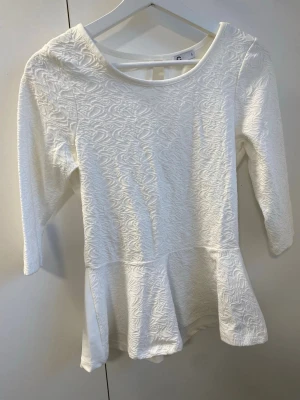 Vit strukturerad topp med peplum - Säljer en vit peplumtop. Toppen har ett snyggt strukturmönster framtill, rund halsringning och trekvartsärm. Modellen är figurnära upptill och har en volang nertill som ger en peplumeffekt. Perfekt att styla med jeans eller kjol.