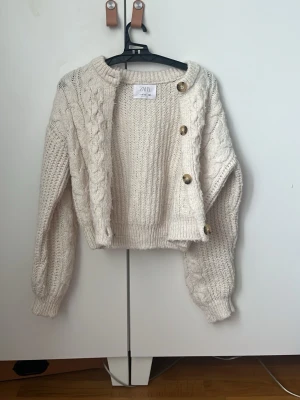Krämvit stickad tröja från Zara - Säljer en krämvit stickad tröja från Zara med grov kabelstickning på ärmarna och ribbade muddar. Tröjan har rund halsringning och dekorativa guldknappar på ena axeln. Perfekt för dig som gillar en mysig och trendig look.