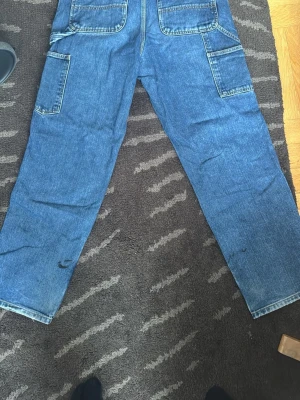 Blå carhartt cargojeans - Säljer ett par blå cargojeans med raka ben och flera stora fickor på sidorna. Jeansen har klassisk passform och är tillverkade i slitstarkt denimtyg. Perfekta för dig som gillar en avslappnad och funktionell stil. Har färgfläckar men kan tas bort med lite möda