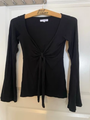 Svart topp från Design by Si - Svart långärmad topp från Design by Si med djup v-ringning och knyt framtill. Modellen är figurnära och har långa, lätt utsvängda ärmar.