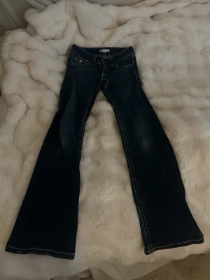 Mörkblå bootcut jeans med kontrastsömmar - Snygga mörkblå bootcut jeans med vita kontrastsömmar och klassisk femficksdesign. Jeansen har låg midja och är tillverkade i ett robust denimtyg som ger en tidlös look. Perfekta för dig som gillar en retroinspirerad stil.