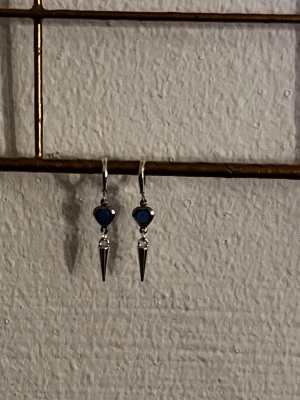 Silvriga örhängen med blå hjärtan - Snygga örhängen i silverfärgad metall med blå hjärtformad detalj och hängande konformad spets. Perfekta för att ge din look en cool och unik touch. Lätta att bära med smidig stängning.
