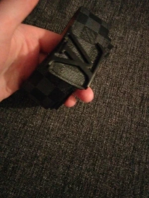 Svart LV-bälte i rutigt läder - Snyggt svart bälte från Louis Vuitton med klassiskt rutigt mönster och stor LV-logga i metall som spänne. Bältet är gjort i läder och har flera hål för justering. Perfekt accessoar för att lyfta din outfit.