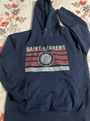 Mörkblå Saint Laurent hoodie - Snygg mörkblå hoodie från Saint Laurent med tryckt logga och ränder i rött och vitt på bröstet. Klassisk känguruficka framtill och justerbar huva med dragsko. Perfekt för en avslappnad och trendig stil.
