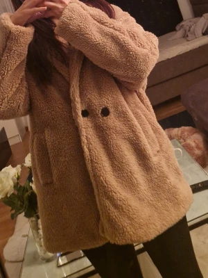 Beige teddyjacka med knappar - Mysig beige teddyjacka med två stora svarta knappar framtill och sidofickor. Jackan har en oversized passform och är tillverkad i fluffigt teddy-material som håller dig varm och stylish. Perfekt för kalla dagar och enkel att matcha med olika outfits.