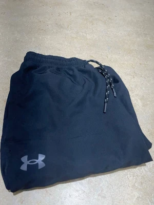Svarta mjukisbyxor från Under Armour - Snygga svarta mjukisbyxor från Under Armour med elastisk midja och snörning. Byxorna har en relaxed passform och är perfekta för chill eller träning. Tillverkade i mjukt och lätt syntetmaterial med logga på benet. 