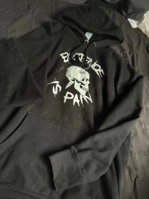 Svart hoodie med dödskalletryck - Svart hoodie med huva och dragsnören, stor vit dödskalle och texten 'EXISTENCE IS PAIN' tryckt på bröstet.
