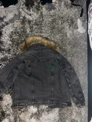 Svart jeansjacka med fuskpäls - Snygg svart jeansjacka med krage i fuskpäls. Jackan har klassisk jeansjacke-design med markerade sömmar och en cool, lite sliten tvätt. Perfekt för dig som vill ha en streetig look med extra edge.