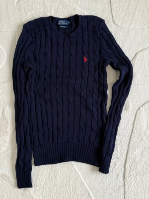 Mörkblå kabelstickad tröja Polo Ralph Lauren -  Helt ny! 2000 kr nypris. Kabelstickad mörkblå tröja från Polo Ralph Lauren med rund halsringning och röd broderad logga på bröstet. Tröjan har långa ärmar och ribbade muddar vid ärmslut och nederkant. Perfekt för en klassisk och stilren look.