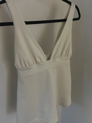 Vit topp med knyt i ryggen - Supersnygg vit omlottopp med djup v-ringning och breda axelband. Toppen har en feminin passform och knyts med en stor rosett i ryggen. Tillverkad i ett mjukt, slätt material som faller fint. Perfekt att styla med jeans eller kjol för en fräsch look.