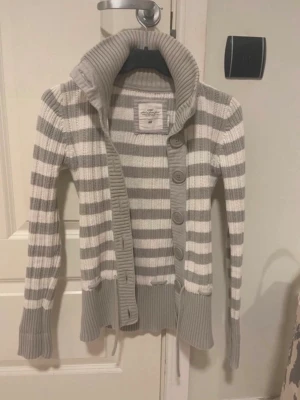 Randig stickad kofta med stor krage - Snygg stickad kofta liknanade Abercrombie & Fitch i grått och vitt med breda ränder. Koftan har stor ribbad krage, knappar framtill och långa ärmar med ribbade muddar. Märket är vintage h&m 