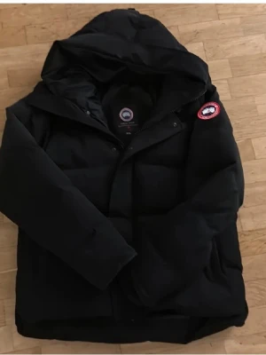 Svart dunjacka från Canada Goose - Säljer en svart dunjacka från Canada Goose med klassisk patch på ärmen och stor huva. Jackan har en puffig design, dragkedja och knappar framtill samt två sidofickor. Perfekt för kalla dagar och riktigt snygg till streetwear-stilen.