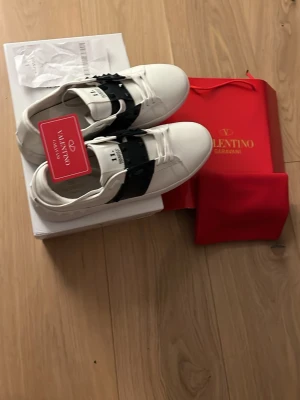 Valentino opens med taggar - Snygga vita sneakers från Valentino Garavani med svart band och nitar över vristen. Skorna har en klassisk rund tå och platt sula. Tillverkade i skinn för en lyxig känsla och stilren look. Perfekta för dig som vill sticka ut med exklusiva detaljer.