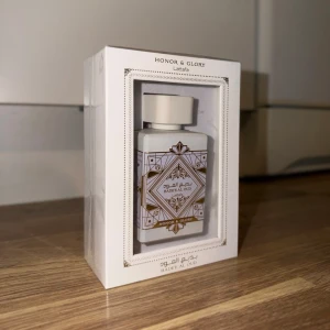 Honor & Glory parfym från Lattafa - Exklusiv parfymflaska från Lattafa i serien Honor & Glory. Flaskan är vit med gulddetaljer och geometriskt mönster, levereras i elegant vit förpackning med transparent framsida. Doften heter Bade'e Al Oud och har en lyxig design som sticker ut i badrumshyllan.