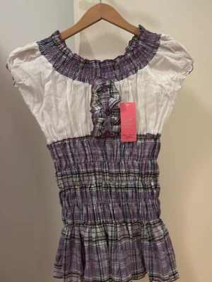 Lila och vit rutig blus - Supersöt blus i lila och vitt med rutigt mönster och smockad midja. Blusen har korta ärmar, volangdetaljer och knappar framtill. Materialet känns som bomull och toppen har en luftig och somrig vibe. Perfekt för dig som gillar en feminin och avslappnad stil.