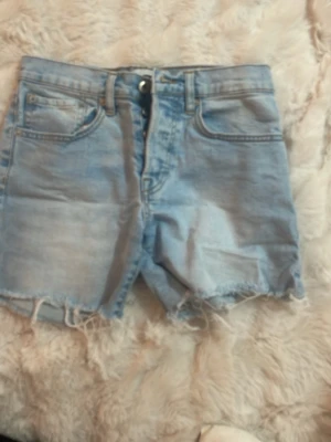 Ljusblå denimshorts med rå kant - Säljer ett par ljusblå denimshorts från Lager 157 med råa kanter och klassisk femficksdesign. Shortsen har hög midja och stängs med knappar. Perfekta för varma dagar och enkel att matcha med olika toppar.
