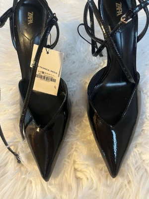 Svarta lackpumps med spetsig tå från Zara - Snygga svarta pumps från Zara i glansigt lackmaterial med spetsig tå och smala remmar som knyts runt ankeln. Klackarna är höga och ger en elegant siluett. Perfekta för dig som vill ha en trendig och stilren look.