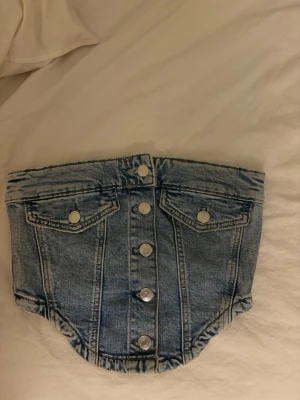 Jeans tubtopp med knappar - Cool tubtopp i ljusblå jeans med silverfärgade knappar framtill och två fejkade bröstfickor. Toppen har en bandeauform och är perfekt för en trendig streetstyle-look. Materialet är klassisk denim och toppen är axelbandslös.