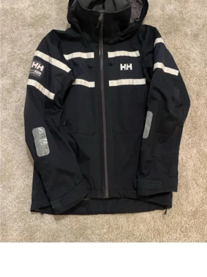 Svart Helly Hansen vindjacka med reflex - Svart vindjacka från Helly Hansen med huva och reflexdetaljer på bröst, armar och rygg. Jackan har dragkedja framtill, två fickor och HH-logga på bröstet. Perfekt för blåsiga dagar och syns bra i mörker tack vare reflexerna.
