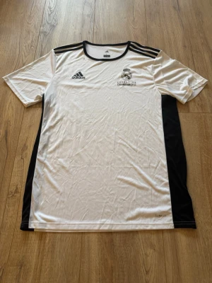 Real Madrid Adidas träningströja - Vit och svart träningströja från Adidas med Real Madrid-logga på bröstet. Tröjan har svarta detaljer på axlar och sidor, samt Adidas klassiska ränder på axlarna. Tillverkad i lätt och ventilerande material, perfekt för fotbollsträning.