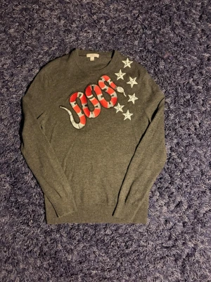 COS tröja - Snygg mörkgrå stickad tröja med broderad röd och vit orm samt sex vita stjärnor på framsidan. Tröjan har rund halsringning och långa ärmar. Materialet är mjukt och tunt, perfekt för lager på lager. Hör av er vid minsta fundering!