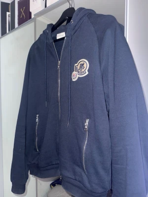 Hoodie - Snygg mörkblå hoodie med dragkedja framtill, två fickor med dragkedja och stor broderad. Hoodien har huva med dragsko och är tillverkad i mjukt bomullsmaterial. Perfekt för en chill och stilren look.