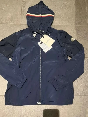 Blå vindjacka från Moncler med huva - HELT NY!! Snygg marinblå vindjacka från Moncler med huva och dragkedja framtill. Jackan har Moncler-logga på ärmen och en randig detalj på huvan i vitt och rött. Tillverkad i lätt polyester, perfekt för blåsiga dagar. Först till kvarn!!