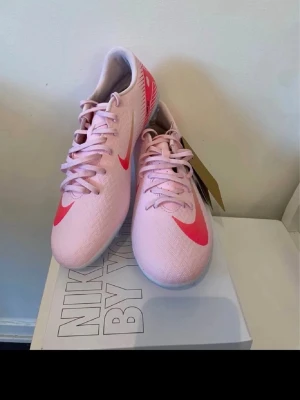 Rosa Nike fotbollsskor med snörning - Säljer ett par snygga rosa Nike fotbollsskor med röda detaljer och klassisk snörning. Skorna har en smal siluett och är tillverkade i syntetmaterial för bästa grepp på planen. Perfekta för dig som vill sticka ut på fotbollsplanen! 40 och 41 storlek