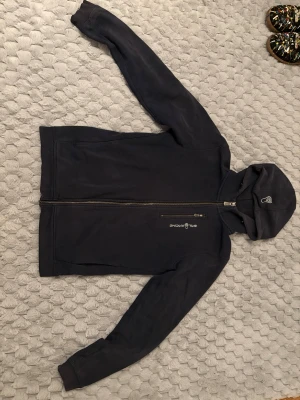 Mörkblå hoodie från sail racing - Snygg mörkblå hoodie från sail racing med dragkedja framtill och huva. Logga på bröstet och en bröstficka med dragkedja. Tillverkad i mjukt material som passar perfekt till chill eller streetwear.