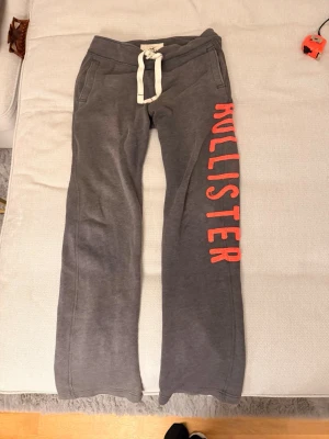 Gråa mjukisbyxor från Hollister - Gråa mjukisbyxor från Hollister med breda ben och vit snörning i midjan. Stora röda bokstäver längs ena benet. Sköna och avslappnade, perfekta för chill dagar. Tillverkade i mjukt bomullsmaterial.