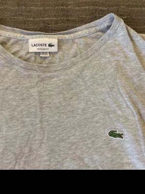 Grå långärmad tröja från Lacoste - Ljusgrå långärmad tröja från Lacoste med klassisk rund halsringning och den ikoniska gröna krokodilloggan broderad på bröstet. Tillverkad i mjukt bomullsmaterial och har en avslappnad regular fit med en bredare form Perfekt för en clean och stilren look.