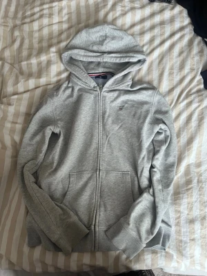 Ljusgrå hoodie från Gant - Säljer en ljusgrå hoodie från Gant med dragkedja framtill och klassisk huva. Hoodien har två fickor, ribbade muddar och en liten Gant-logga broderad på bröstet. Materialet är mjukt och skönt, perfekt för chill dagar. Snygg detalj med randig insida på kragen.