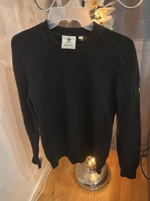 Svart stickad tröja från Lyle & Scott - Snygg svart stickad tröja från Lyle & Scott med rund halsringning och ribbade detaljer på axlarna. Tröjan har lång ärm och en diskret logotyp på ärmen. Perfekt för en stilren och enkel look.