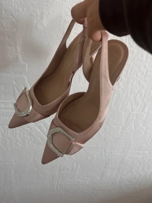 Rosa pumps med strassdetalj - Snygga ljusrosa pumps i satin med spetsig tå och låg klack. Skorna har öppen häl och dekoreras med en stor strassprydd detalj framtill. Perfekta för dig som vill ha en elegant och trendig look. Skriv till mig för intresse! 