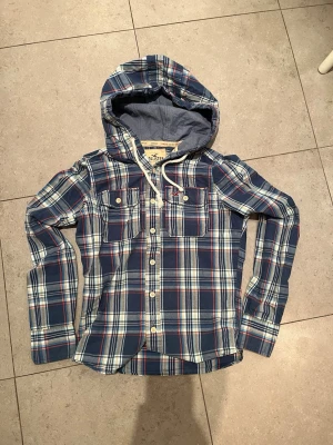 Sällsynt hollister tröja - Snygg blårutig skjorttröja från Hollister med huva och knappar framtill. Tröjan har två bröstfickor med lock och vita knappar. Materialet är mjukt och bekvämt, perfekt för en avslappnad stil.