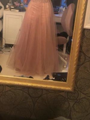 Lång rosa balklänning med spetsliv - Elegant långklänning i ljusrosa med ett detaljerat spetsliv och voluminös tyllkjol. Klänningen har smala axelband och en vacker, svepande siluett som passar perfekt till bal eller fest. Materialet är troligtvis polyester och tyll. Köpt för 1200 kr.