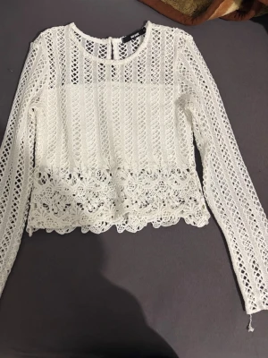 Vit spetsblus med lång ärm - Superfin vit blus i spets med långärmad design och rund halsringning. Blusen har vackra spetsdetaljer och en lätt croppad passform med dekorativ kant nertill. Perfekt för dig som gillar romantisk och trendig stil.