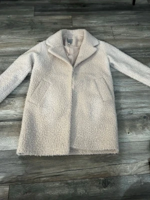 Beige teddyjacka med krage - Mysig beige teddyjacka med klassisk krage och två snedställda fickor framtill. Jackan har en rak passform och stängs med dolda knappar. Perfekt för dig som vill ha en stilren och fluffig look.