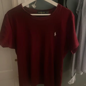 Vinröd t-shirt från Ralph Lauren - SSnygg vinröd t-shirt från Ralph Lauren med klassisk passform och rund hals. T-shirten har korta ärmar och den ikoniska broderade loggan på bröstet. Tillverkad i mjuk bomull som känns skön mot huden. Storlek l passar som m 