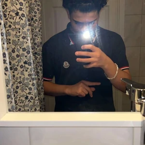 Svart Moncler t-shirt med röd/vita detaljer - Svart t-shirt från Moncler med klassisk logga på bröstet. T-shirten har korta ärmar och krage med röd och vit randig detalj. Passformen är normal och materialet känns som mjuk bomull. Perfekt för dig som gillar stilrena märkesplagg.