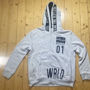 Ljusgrå hoodie från Zara Boys - Ljusgrå hoodie med dragkedja från Zara Boys. Tryck med texten 'Young and Restless' på huvan och 'WRLD TOUR 01' framtill. Mjuk bomullskänsla, fickor på sidorna och långa ärmar. Perfekt för en avslappnad streetstil.
