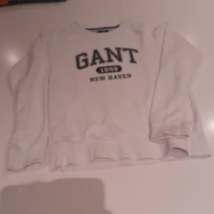 Vit sweatshirt från GANT, 158/164 - Vit sweatshirt från GANT med tryck 'GANT 1949 NEW HAVEN' på bröstet. Klassisk rund halsringning och långa ärmar. Perfekt för dig som gillar en clean och stilren look. Mjuk och skön, passar till jeans eller mjukisbyxor.