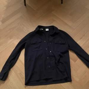 Mörkblå overshirt från Selected Homme - Stilren mörkblå overshirt från Selected Homme med klassisk krage, två bröstfickor med lock och tryckknappar framtill. Skjortan är långärmad och har en rak passform, perfekt att bära öppen eller stängd. Materialet är bomull för en skön känsla.